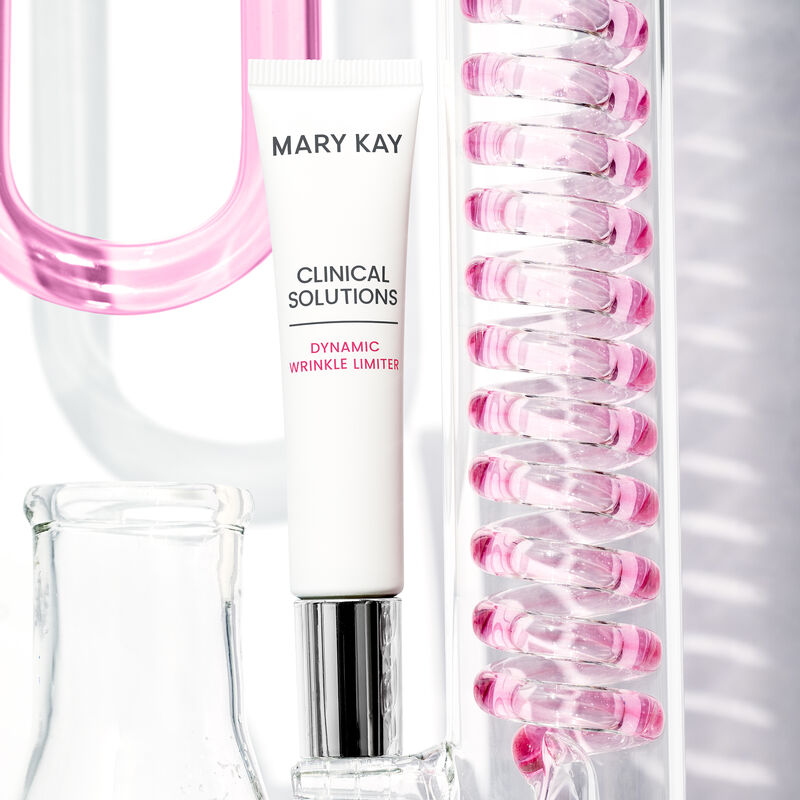 Tube of Mary Kay Clinical Solutions Dynamic Wrinkle Limiter standing on its cap surrounded by glass laboratory containers filled with pink liquid